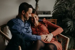 photographe grossesse de parents installés dans un fauteuil de leur salon qui rient en regardant le ventre rond