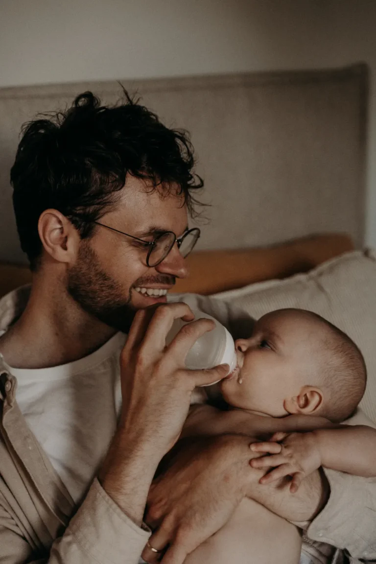 un père assis sur son lit sourit en donnant le biberon à son bébé de 9 mois dans ses bras, photographié par une photographe bébé