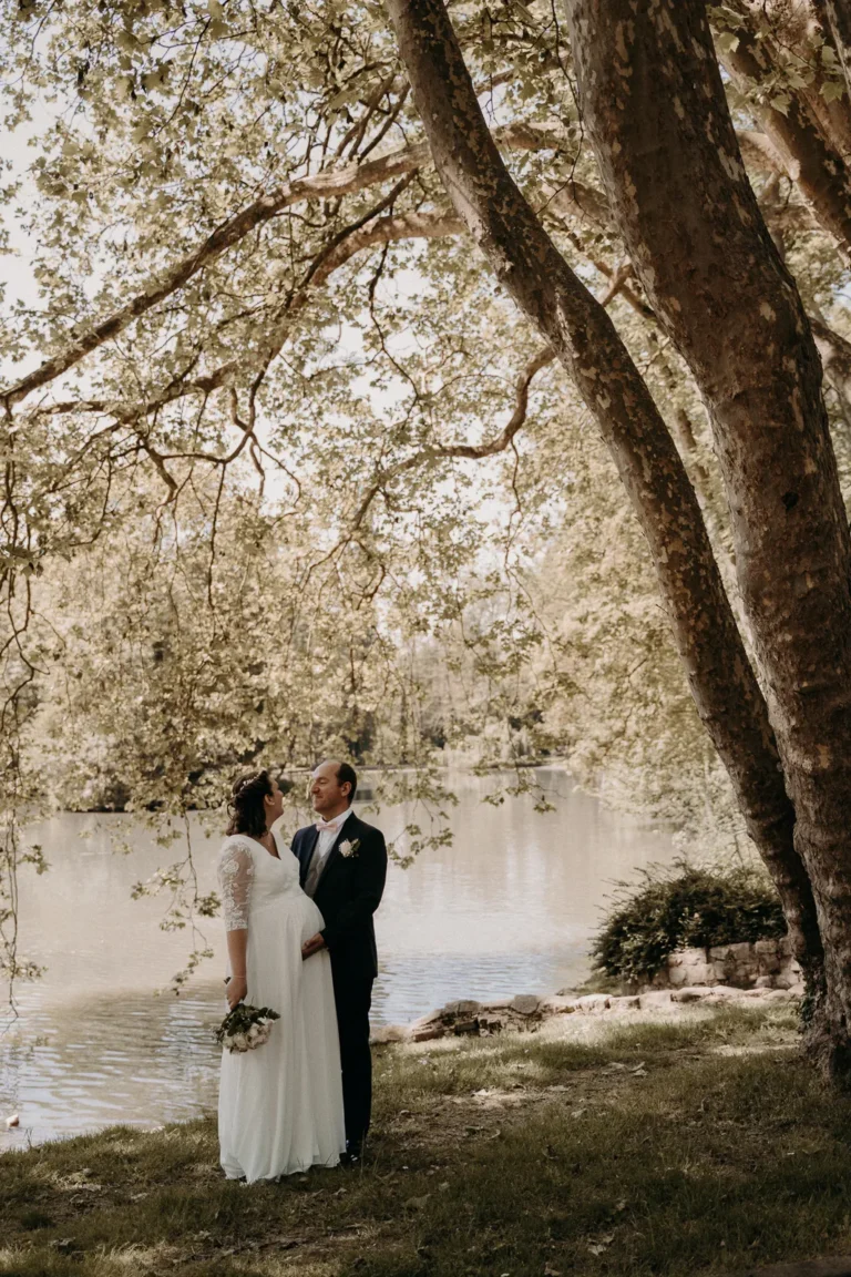 mariée enceinte et son mari se regardent avec amour au bord d'un lac sous un arbre photographiés par une photographe de mariage